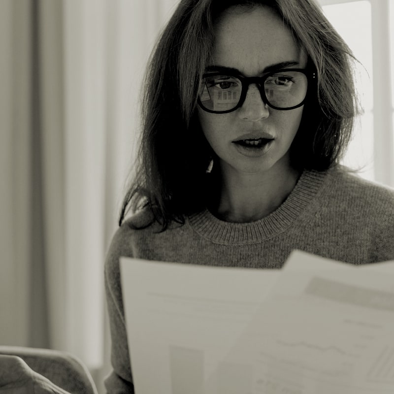 Black and white image of a caucasian woman looking shocked at the papers she's holding in her hands
