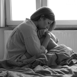 Black and white image of a caucasian woman sitting in bed next to a window looking somber