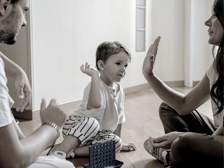 A young child celebrates a successful play while sitting on the floor between two adults, possibly during a board game, a scene reminiscent of happier times before discussions about divorce mediation.