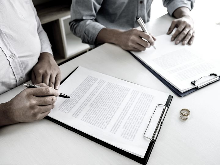 Two individuals seated at a table during a divorce document signing, with a wedding band prominently displayed.